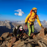 Three roped climbers wearing helmets ascend a rocky alpine ridge, with snow-covered peaks behind them.