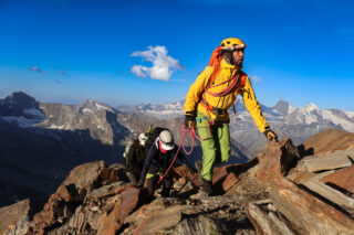 Three roped climbers wearing helmets ascend a rocky alpine ridge, with snow-covered peaks behind them.