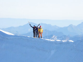 Three roped climbers on a snowy alpine ridge, with one raising an ice axe and mountain peaks in the distance.
