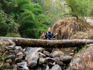 Trekker sitting with legs crossed on a log above a rocky stream surrounded by bamboo and forest.