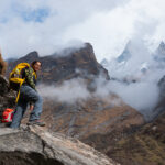 Trekker with a yellow backpack on rocky steps, with cloud and snowy mountain peaks behind.