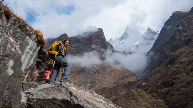 Trekker with a yellow backpack on rocky steps, with cloud and snowy mountain peaks behind.