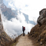 Trekker with a red backpack on a narrow mountain trail between steep cliffs and low cloud.