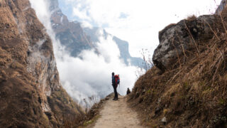 Trekker with a red backpack on a narrow mountain trail between steep cliffs and low cloud.