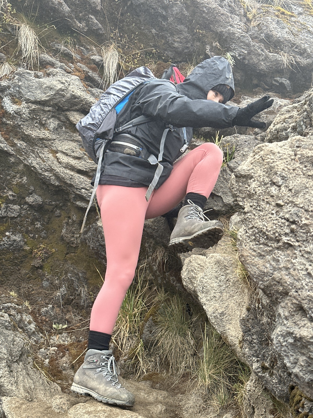A trekker in pink trousers lifts a boot onto a higher rock while a gloved hand touches the rock beside them.
