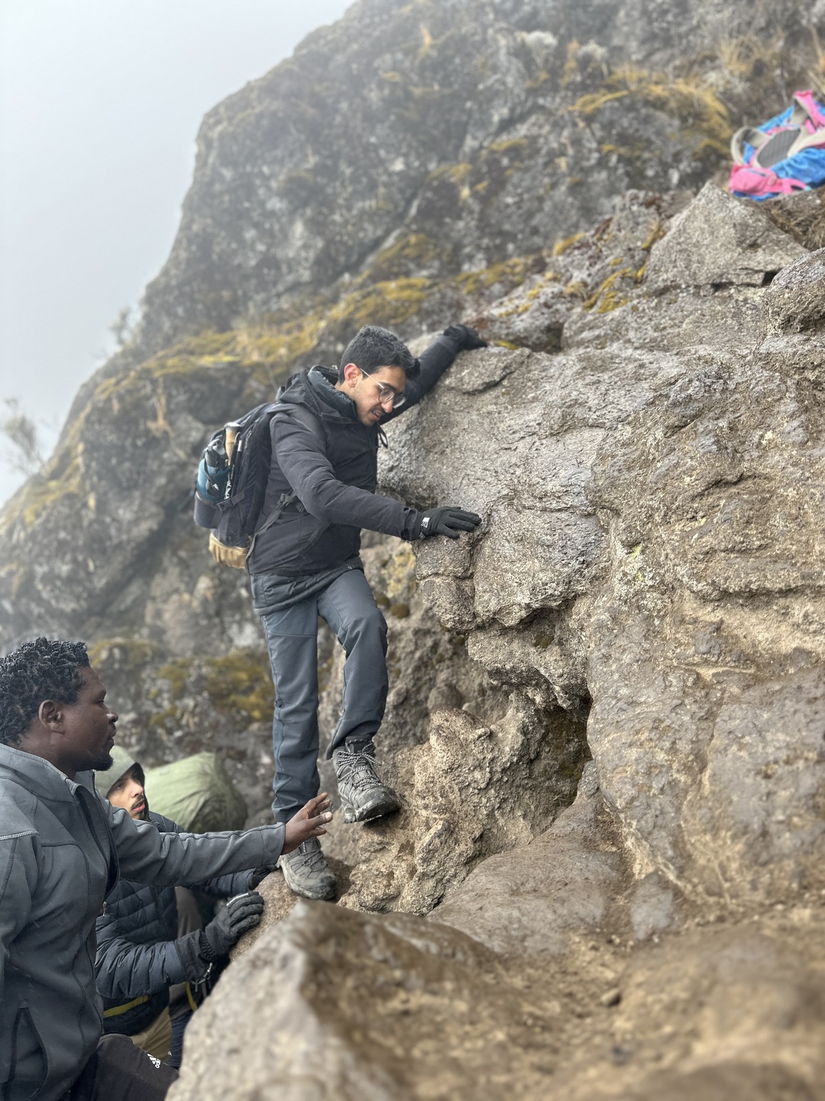 A trekker stands on narrow rock footholds with both hands against the rock face; two people are below and the slope recedes into mist.