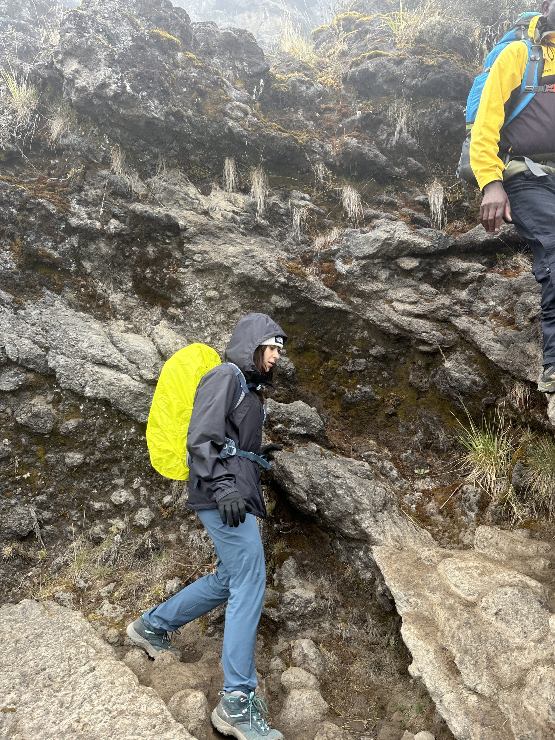 A trekker in blue trousers steps between rocks on the Barranco Wall, with another person above to the right.