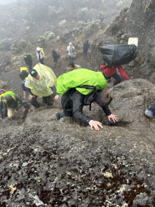 Viewed from above, a trekker with a bright green pack cover uses both hands on a rock step, with several people below in mist.