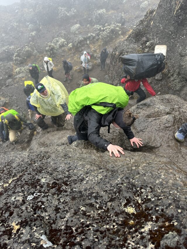 Viewed from above, a trekker with a bright green pack cover uses both hands on a rock step, with several people below in mist.