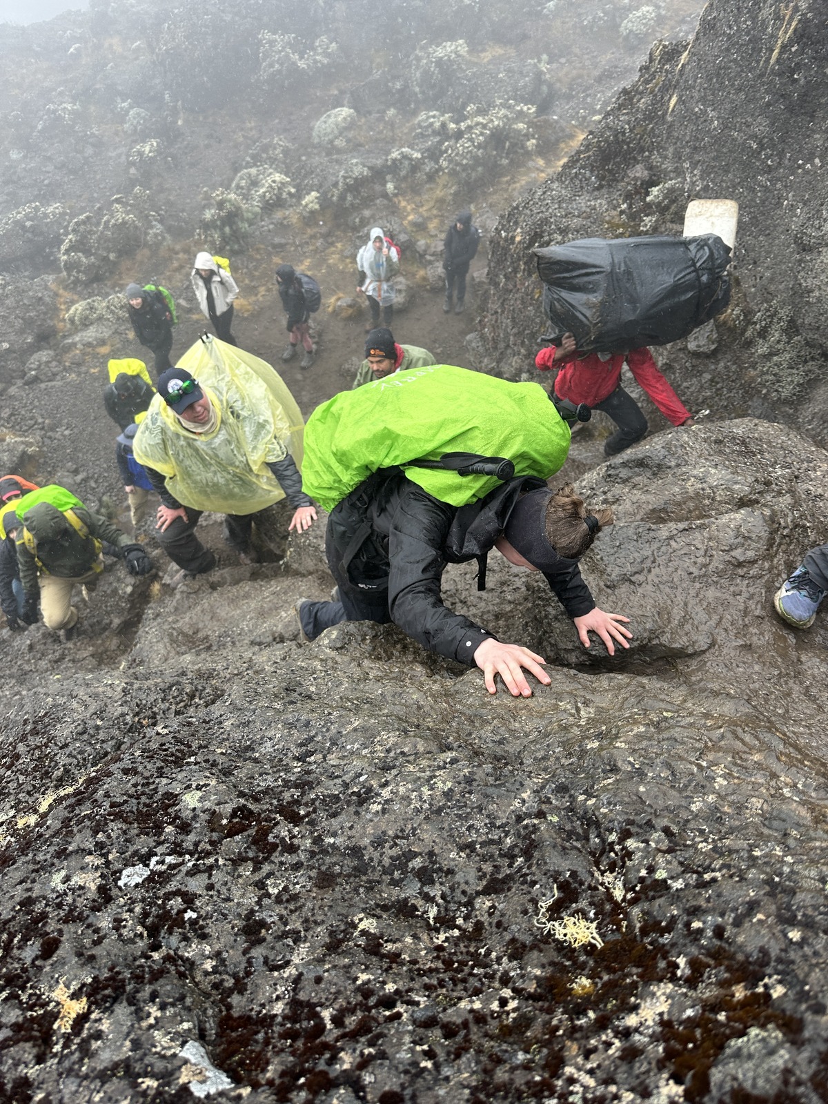 Viewed from above, a trekker with a bright green pack cover uses both hands on a rock step, with several people below in mist.