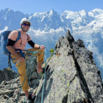 Rami Rasamny wearing a helmet, harness and blue backpack on a rocky ridge, with snowy mountains behind.
