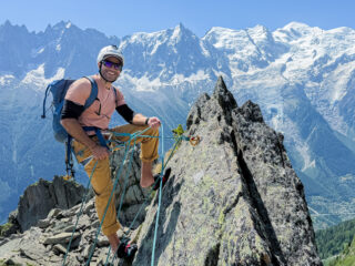 Rami Rasamny wearing a helmet, harness and blue backpack on a rocky ridge, with snowy mountains behind.