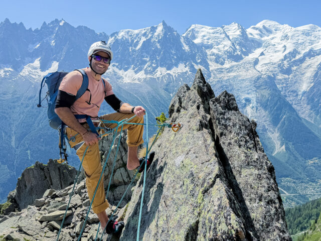 Rami Rasamny wearing a helmet, harness and blue backpack on a rocky ridge, with snowy mountains behind.