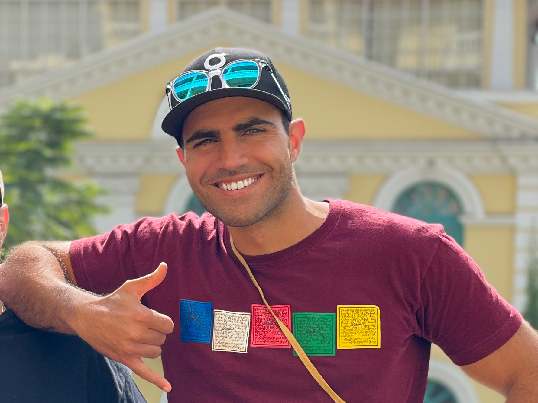 Rami Rasamny smiling outdoors in a burgundy T-shirt and cap, with sunglasses resting on the brim.