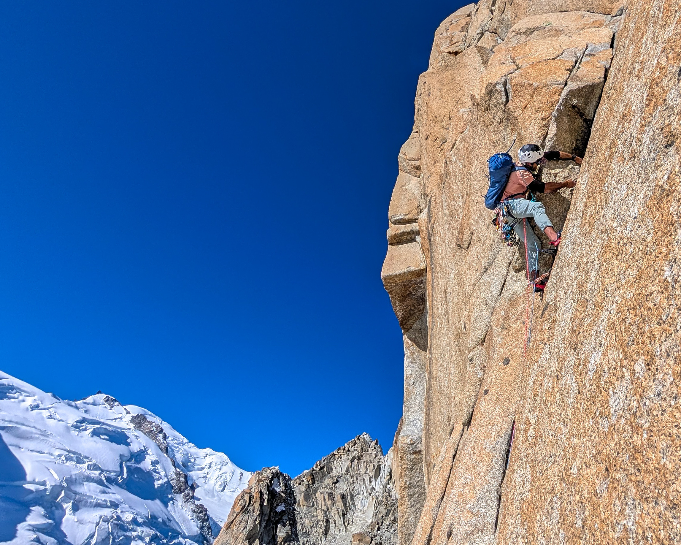 A roped climber wearing a helmet and blue backpack ascends a steep granite face, with snowy mountains behind.