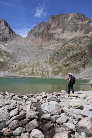Hiker beside a sunlit alpine lake beneath rocky peaks and blue sky on the Tour du Mont Blanc